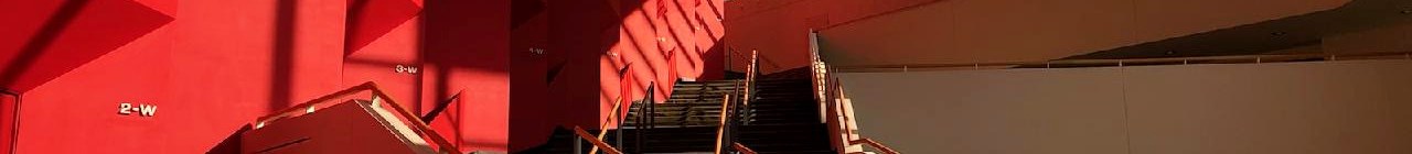 Red multi-angular wall with linear shadows abuts an ascending staircase flanked by a long skylight